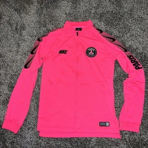 Rosa träningsjacka från Nike - Säljer en snygg rosa träningsjacka från Nike med PSG-logga. Jackan har långa ärmar och dragkedja framtill. Perfekt för träning eller som en färgglad vardagsjacka. 