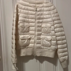 Vit dunjacka från Moncler - Säljer en snygg benvit dunjacka från Moncler i nyskick. Jackan har en quiltad design med dragkedja och två praktiska fickor framtill. Perfekt för våren och sommaren. Den är både stilren och varm, vilket gör den till ett måste i garderoben!