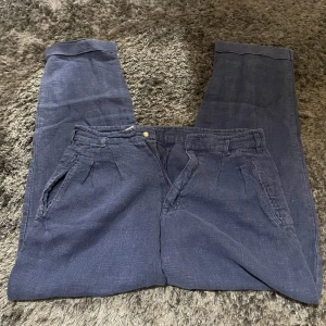 Blå linnebyxor från Natural - Säljer ett par snygga blå linnebyxor från Natural i storlek 48. De är perfekta för sommaren med sin lätta och luftiga känsla. Byxorna har en klassisk design med fickor fram och en knappdetalj bak. Perfekta för både vardag och fest!