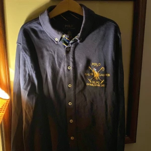 Mörkblå pikétröja från Ralph Lauren - Säljer en snygg mörkblå pikétröja från Ralph Lauren med gul broderad logga på bröstet. Tröjan har knappar framtill och en klassisk krage. Perfekt för en stilren look. Obs det är en barntröja som passar mellan 10-12 år. 