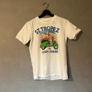 Vit St. Tropez t-shirt med Vespa-tryck - Säljer en vit t-shirt med ett coolt St. Tropez-tryck och en grön Vespa på framsidan. Perfekt för sommardagar och avslappnade utflykter. T-shirten är kortärmad och gjord i mjuk bomull. Passar bra till jeans eller shorts för en chill look.
