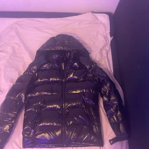 Svart glansig dunjacka från Moncler - Säljer en snygg svart glansig dunjacka från Moncler. Jackan har en dragkedja framtill och en huva för extra värme. Perfekt för kalla vinterdagar. Den är i bra skick och redo för nya äventyr! 🖤