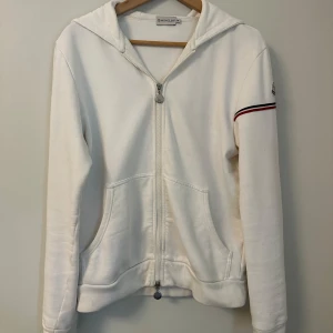 Vit hoodie från Moncler - Säljer en snygg vit hoodie från Moncler i mycket bra skick. Den har en dragkedja framtill och en cool randig detalj i svart och rött på ärmen. Perfekt för både vår och höst! 🖤❤️