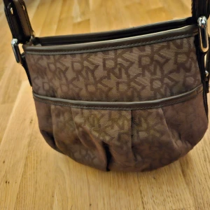 Brun axelväska från DKNY - Snygg brun axelväska från DKNY med monogrammönster. Väskan har en praktisk dragkedja och justerbar axelrem. Perfekt för vardagsbruk och rymmer det mesta du behöver ha med dig. Den är i mycket bra skick och redo för nya äventyr!