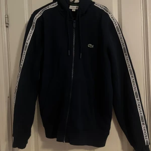 Mörkblå hoodie från Lacoste - Säljer en fin mörkblå hoodie från Lacoste i klassisk passform. Den har en dragkedja framtill och vita detaljer längs ärmarna med Lacoste-logga. Perfekt för höst och vår, och den är superbekväm att ha på sig. Den är i storlek M och är köpt från Zalando, har använt 2-3 gånger och är i mycket bra skick, om det är andra frågor du undrar över Är det bara o hojta till! Obs det är inte fast pris, det går även att diskutera.