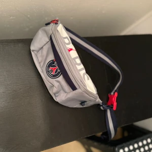 Grå midjeväska från Paris Saint-Germain - Säljer en snygg grå midjeväska från Paris Saint-Germain med coola detaljer. Väskan har en justerbar rem i blått och vitt med en röd spänne. Perfekt för att bära dina småsaker med stil. Den har en dragkedja för säker förvaring och en logga på framsidan.