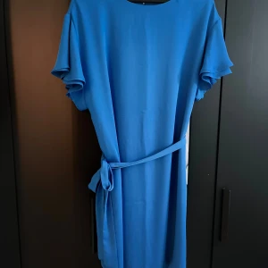 Blå klänning med knytband - Säljer en superfin blå klänning med kortärmad design och ett matchande knytband i midjan. Perfekt för både vardag och festliga tillfällen. Klänningen har en enkel och stilren look som gör den lätt att styla med olika accessoarer.
