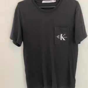 Svart t-shirt från Calvin Klein Jeans - Säljer en stilren svart t-shirt från Calvin Klein Jeans i storlek M. Den har en liten bröstficka med CK-loggan tryckt i vitt. Perfekt för en casual look eller att ha under en jacka. T-shirten är kortärmad och gjord i mjuk bomull. Passar både till vardags och fest!