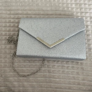 Glittrig silver kuvertväska - Säljer en superfin glittrig kuvertväska i silver. Den har en elegant kedja som axelrem och en snygg V-formad detalj på locket. Perfekt för fest eller nyår! ✨