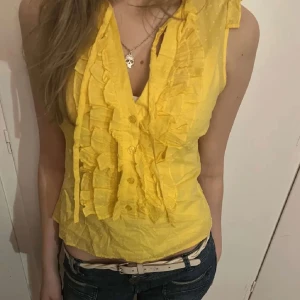 Gul blus med volangdetaljer - Säljer en superfin gul blus med volangdetaljer framtill. Den är ärmlös och perfekt för sommardagar. Blusen har en v-ringad design och är gjord i ett lätt och bekvämt material. Passar perfekt till både jeans och kjol!