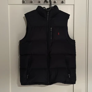  Väst från Polo Ralph Lauren - Säljer en stilren mörkblå dunväst från Polo Ralph Lauren. Västen har en dragkedja framtill och två praktiska fickor med dragkedjor. Perfekt för kyligare dagar när du vill hålla stilen enkel och snygg. Passar om du är ca 160-170cm lång, vid fler frågor eller funderingar är det bara att höra av sig. MVH Grischoutlet.