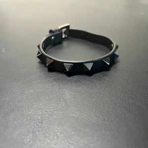 Valentino armband( Rockstud leather bracelet with Ruthenium studs) - Snyggt svart läderarmband från Valentino med rockiga ruteniumnitar. Perfekt för att ge din stil en edgy touch. Armbandet kommer med en praktisk förvaringspåse.