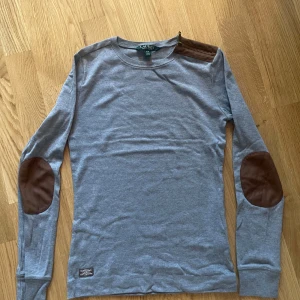 Grå långärmad tröja från Ralph Lauren - Säljer en stilren grå långärmad tröja från Ralph Lauren med bruna armbågslappar och en brun axeldetalj. Perfekt för en avslappnad och snygg look. Tröjan är i mjukt material och har en normal passform.