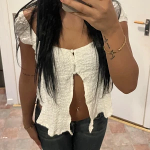 Vit offshoulder topp - Säljer en söt vit offshoulder topp med knappar framtill. Toppen har en lätt rynkad textur och är perfekt för varma dagar. Den är kort och ger en avslappnad look.