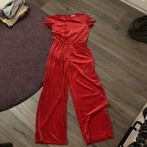 Röd byxdress från KappAhl - Snygg röd byxdress från KappAhl i ett glansigt material med ribbad struktur. Den har korta ärmar och en elastisk midja för en bekväm passform. Nyskick bara använd 2 gånger.