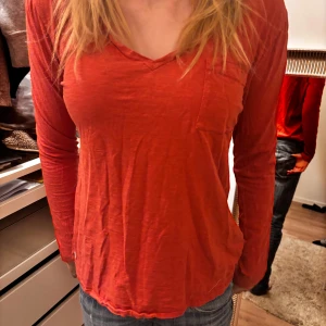Röd långärmad topp med v-ringning - Säljer en snygg röd långärmad topp med v-ringning och en liten bröstficka. Perfekt för en avslappnad stil. Passar bra till jeans eller kjol.