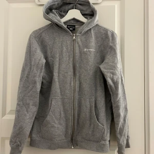 Grå hoodie från Champion - Säljer en klassisk grå hoodie från Champion med dragkedja och huva. Tröjan har långa ärmar och två praktiska fickor framtill. Perfekt för en avslappnad stil. 🩶
