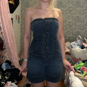 jeans jumpsuit - Trendig axelbandslös jeans jumpsuit med knappar framtill. Den har en figurnära passform och praktiska fickor. Perfekt för en avslappnad stil med en touch av edge.