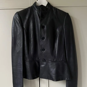 Ralph Lauren-jacka i perfekt skick - Snygg mörkblå, nästan svart skinnjacka från Ralph Lauren med knappar framtill. Jackan har en stilren design med långa ärmar, är figursydd och har hög krage. Perfekt för att ge din outfit en edgy touch. 🖤Mjuk lammnappa med innerfoder. Använd fåtal gånger och i perfekt skick! US-storlek 4, d v s storlek 32-34 i svensk storlek. Nypris ca 15000kr.