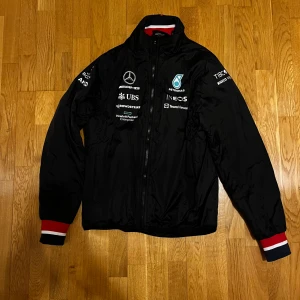 Svart jacka från Mercedes AMG Petronas - Säljer en svart jacka från Mercedes AMG Petronas Formula One Team. Jackan har broderade logotyper och detaljer i rött, vitt och blått. Den har en dragkedja framtill och långa ärmar med ribbade muddar. Perfekt för racingfans! 🏎️