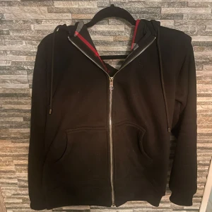 Svart hoodie från Burberry - Säljer en svart hoodie från Burberry med dragkedja och klassiskt rutigt mönster i huvan. Pris kan diskuteras