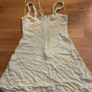 Gul och vit klänning med blommönster - Charmig gul och vit klänning med blommönster och smala axelband. Klänningen har en söt snörning framtill och en lätt, luftig design som passar perfekt för varma dagar.