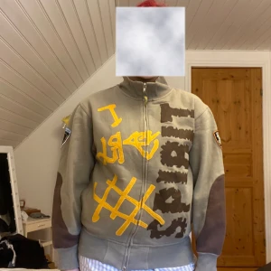 Green jumper from SWAG  - Säljer en grön jumper från SWAG med ett iögonfallande gult grafiskt tryck på framsidan. Jumper har långa ärmar med bruna detaljer och en dragkedja framtill. Den har även ett märke på ärmen som ger en unik touch. DM att prata om priset!