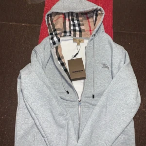 Grå huvtröja från Burberry - Säljer en stilren grå huvtröja från Burberry med klassiskt rutigt mönster i huvan. Tröjan har dragkedja och en broderad logga på bröstet. Perfekt för en avslappnad och trendig look.