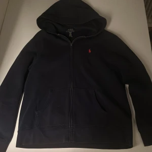 Mörkblå hoodie från Ralph Lauren - Säljer en stilren mörkblå hoodie från Ralph Lauren med dragkedja och huva. Den har en liten röd logga på bröstet och praktiska fickor framtill. Perfekt för en avslappnad look.
