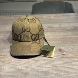 Gucci jumbo beige helt ny med tags pris kan disskuteras  - Snygg beige keps från Gucci med det ikoniska GG-mönstret. Kepsen har en justerbar rem baktill i brunt läder och är tillverkad i ett slitstarkt material. Perfekt för att ge din outfit en lyxig touch. Dustbag tillkommer!