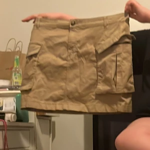 Beige kortkjol med fickor - Säljer en beige kortkjol med praktiska fickor på sidorna. Kjolen har en knapp och dragkedja framtill för enkel påklädning. Perfekt för en avslappnad stil.