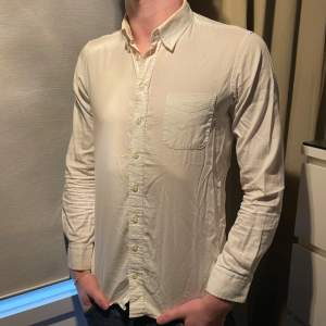 Säljer en stilren vit skjorta från GANT med klassisk krage och bröstficka. Skjortan har långa ärmar och knappar framtill. Perfekt för en snygg och avslappnad look. Skjotan har blivit lite gul i kragen därav priset