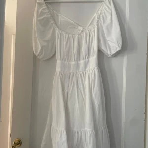 Vit klänning med puffärmar - Charmig vit klänning med puffiga kortärmar och en söt knytning i ryggen. Klänningen har en luftig passform och är perfekt för varma dagar. Den har en romantisk och tidlös stil som passar många tillfällen. Passar bra till studenten