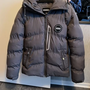 Grå pufferjacka från Hoodrich - Säljer en grå pufferjacka från Hoodrich med tryck på luvan och ryggen
