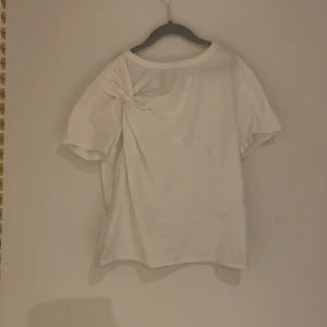 Vit kortärmad topp från H&M Divided - Säljer en stilren vit kortärmad topp från H&M Divided. Toppen har en unik knutdetalj på framsidan som ger en extra touch. Perfekt för en enkel och chic look. Passar bra till både jeans och kjol.