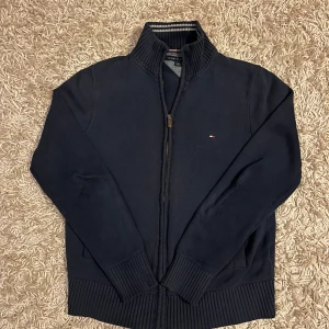 Mörkblå finstickad tröja med dragkedja från Tommy Hilfiger - Säljer en mörkblå finstickad tröja från Tommy Hilfiger med dragkedja och ribbad krage. Tröjan har ett litet broderat logotypmärke på bröstet och ribbade muddar vid ärmslut och nederkant. Perfekt för en stilren look. Har en liten skavank vid ena armen som syns på bild men inte när man bär den, övrigt fint skick. Fraktas antigen samma dag som köp eller dagen därpå💯🙌