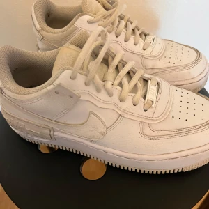 Vita Nike sneakers - Säljer ett par klassiska vita Nike sneakers med snörning och perforerade detaljer på ovansidan. Skorna har en robust sula och den ikoniska Nike-loggan på sidan och hälen. Perfekta för en stilren look.