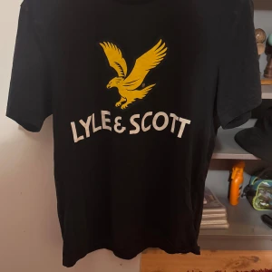 Svart t-shirt från Lyle & Scott - Snygg svart t-shirt från Lyle & Scott med deras ikoniska gula örn och logga på bröstet. Perfekt för en stilren och avslappnad look.