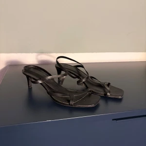 Svarta sandaler med klack - Snygga svarta sandaler med smala remmar och klack. Perfekta för en stilren look. De har en elegant design med en spetsig tå och tunna remmar som korsar över foten.