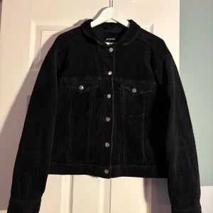 Svart manchesterjacka från Monki - Snygg svart manchesterjacka från Monki med knappar framtill. Jackan har en klassisk design med två bröstfickor och långa ärmar. Perfekt för en stilren look.