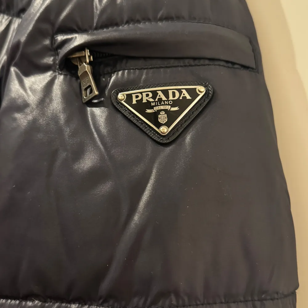 Nypris ca: 16000kr Säljer en mörkplå pufferjacka från Prada med dragkedja och tryckknappar. Jackan har en klassisk design med långärmade ärmar och en hög krage. Den är tillverkad i ett glansigt nylon och har Pradas logotyp på ärmen. Perfekt för kyliga dagar.. Takit.