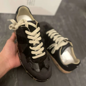 Maison Margiela - Snygga svarta sneakers med beige snörning och en unik sula med stänkmönster. Skorna har en stilren design med en kombination av olika material som ger en cool look.