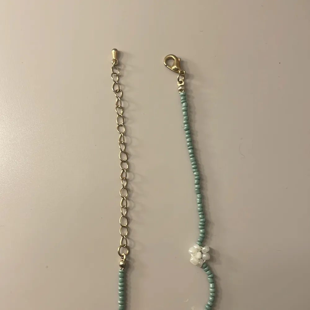 Halsband med turkosa små pärlor och vita blommor av pärlor. Halsbandet har en justerbar kedja i guldton och ett karbinlås. Perfekt för att ge en färgglad touch till din outfit.. Asusteet.