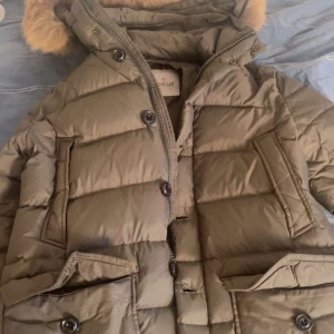 Olivgrön dunjacka från Moncler - Säljer en snygg olivgrön dunjacka från Moncler med pälskantad huva. Jackan har en quiltad design med både dragkedja och knappar framtill. Den har praktiska fickor och en cool serietidningsdetalj på insidan. Perfekt för kyliga dagar! Äkta såklart, inget kvitto dock