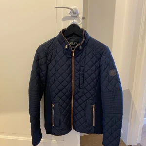 Mörkblå quiltad jacka från Hollies - Snygg mörkblå quiltad jacka från Hollies med dragkedja och knappar. Jackan har en stilren design med långärmade ärmar och två praktiska fickor med dragkedja framtill. Perfekt för kyligare dagar.
