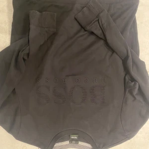 Svart tröja från Hugo Boss - Säljer en stilren svart tröja från Hugo Boss med subtilt broderad logga på framsidan. Tröjan har långa ärmar och ribbade muddar vid ärmslut och nederkant. Perfekt för en elegant och avslappnad look. Riktigt snygg 