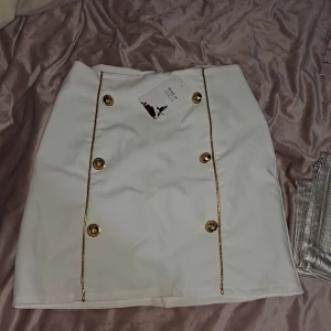 Vit kjol med guldknappar - Snygg vit kjol med dekorativa guldknappar och dragkedja bak. Tillverkad i Italien, storlek 8. Perfekt för en stilren look.