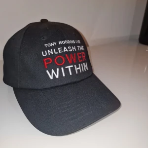 Tony Robbins keps - Tony Robbins keps 'UNLEASH THE POWER WITHIN' broderad i vitt och rött framtill. Baktill finns texten 'Breakthrough Success Resources' i blått och vitt. Kepsen är tillverkad i 100% bomull och har justerbar passform. Garderobsrensning så kika gärna på övriga auktioner.