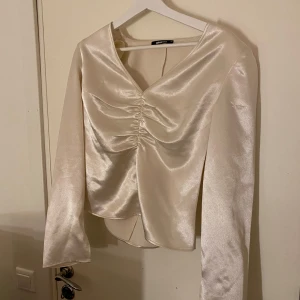 Vit satinblus från Gina Tricot - Elegant vit blus i satin från Gina Tricot med långa ärmar och v-ringad design. Blusen har en snygg rynkdetalj framtill som ger en stilfull touch. 