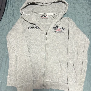 Grå hoodie från Saint Vincent - Säljer en grå hoodie från Saint Vincent med dragkedja och tryck på bröstet. Perfekt för en avslappnad stil. Huvan ger extra mysfaktor och den är lätt att matcha med andra plagg.